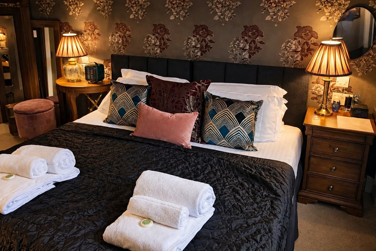 Luxury hotel bedroom with black bed, white rolled towels, patterned wallpaper, warm lighting, and wood nightstands
