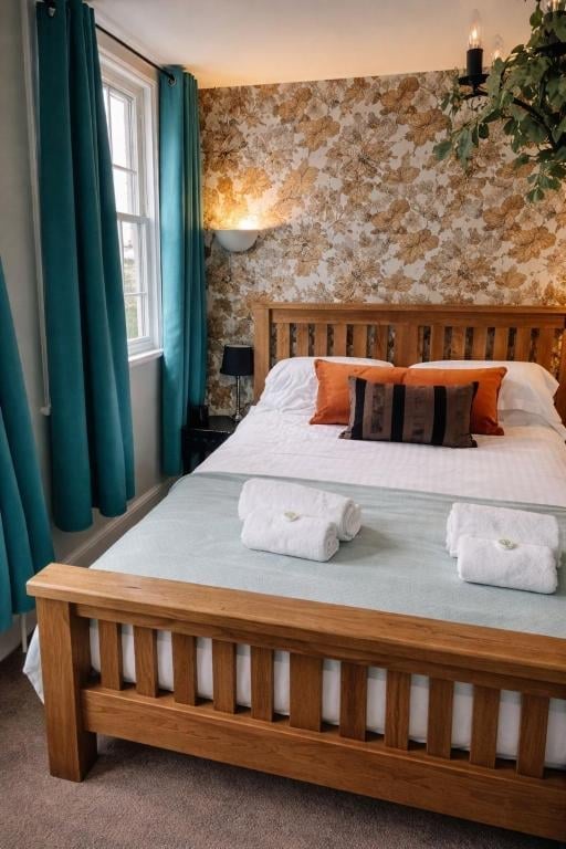 A Union Inn bedroom with a wooden bed, white bedding, orange and striped accent pillows, gold patterned wallpaper and teal curtains