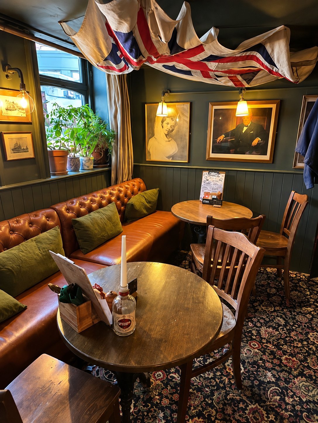 British pub interior with brown leather booth seating, round wooden tables, framed portraits on dark walls, and a British flag overhead